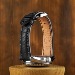 Pode incluir: Uma pulseira de relógio de couro preto com uma fivela prateada. A pulseira está em um suporte de madeira e tem o texto "Badalassi Carlo Leather Goods" impresso na parte de trás.