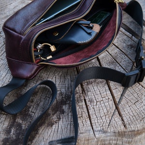 May include: A brown leather bum bag with a black adjustable strap. The bag is open and shows a variety of items inside, including a phone, wallet, and keys.