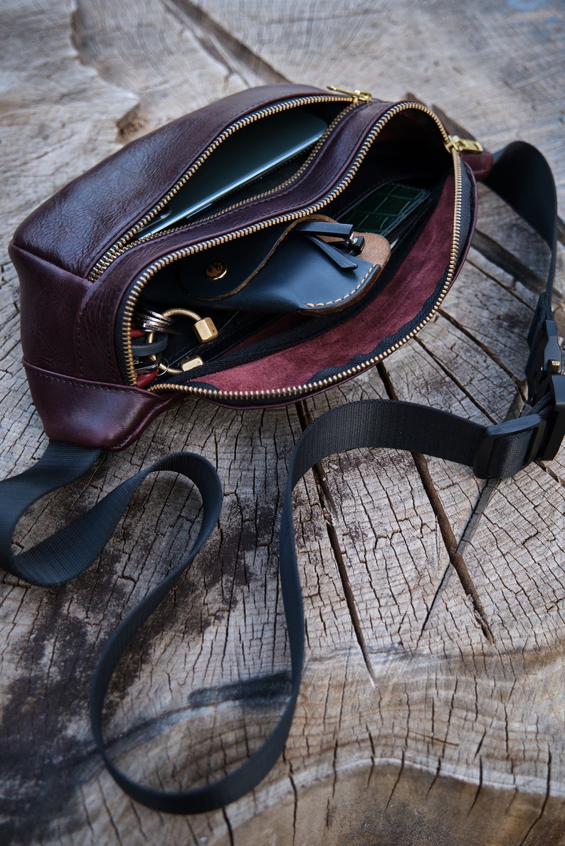 May include: A brown leather bum bag with a black adjustable strap. The bag is open and shows a variety of items inside, including a phone, wallet, and keys.