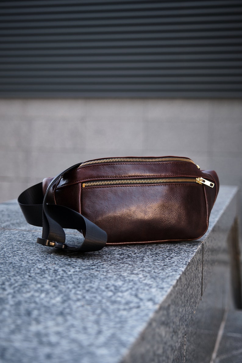 May include: A brown leather bum bag with a black strap and gold zips. The bag is sitting on a grey stone surface.