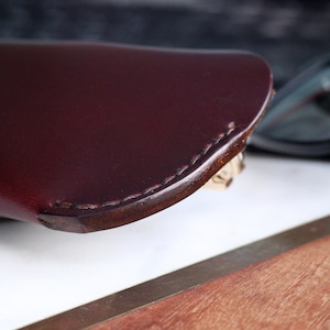 May include: A close-up of a burgundy leather glasses case with a stitched edge. The case is resting on a wooden surface.