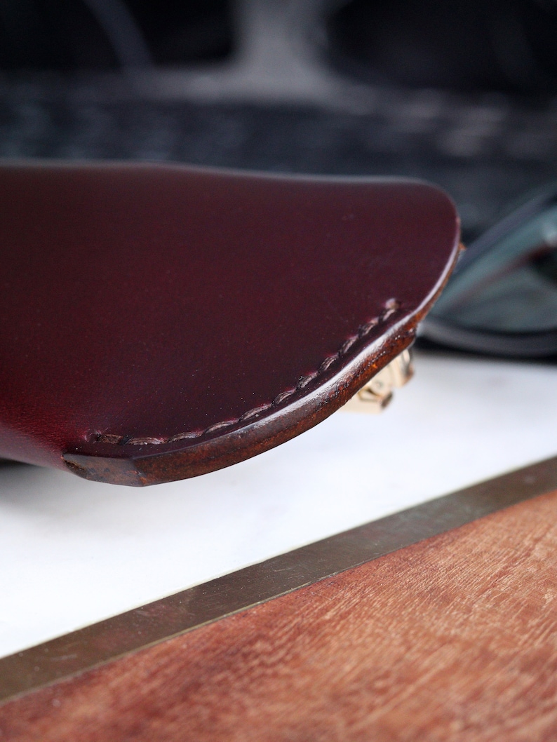 May include: A close-up of a burgundy leather glasses case with a stitched edge. The case is resting on a wooden surface.