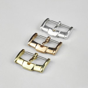 Pode incluir: Três fivelas de relógio em tons de ouro, ouro rosa e prata. Cada fivela tem uma moldura retangular com uma parte superior curva e uma barra de metal para prender uma pulseira de relógio. As fivelas são exibidas em uma superfície branca.