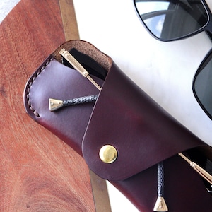 May include: A burgundy leather glasses case with a gold button closure and a black cord. The case is folded over and a pair of black sunglasses are inside.