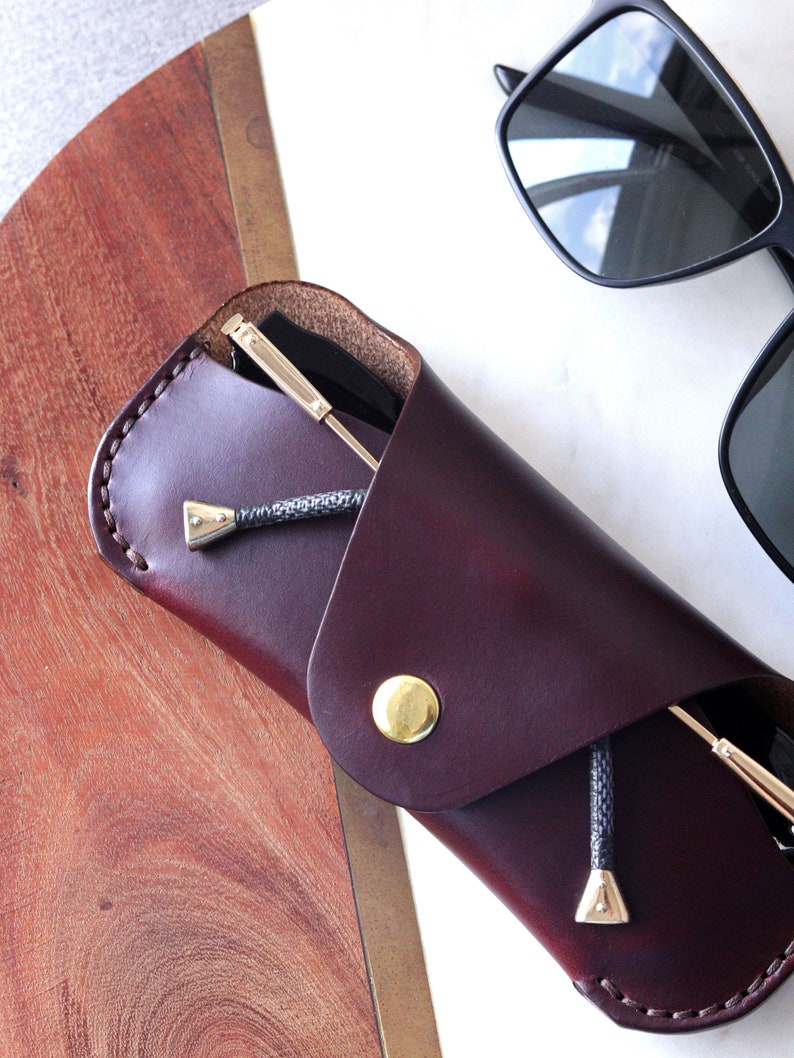 May include: A burgundy leather glasses case with a gold button closure and a black cord. The case is folded over and a pair of black sunglasses are inside.