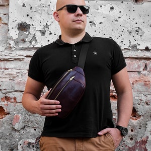 May include: A person wearing a black polo shirt with a brown leather fanny pack worn across the chest. The person is wearing brown trousers and sunglasses.