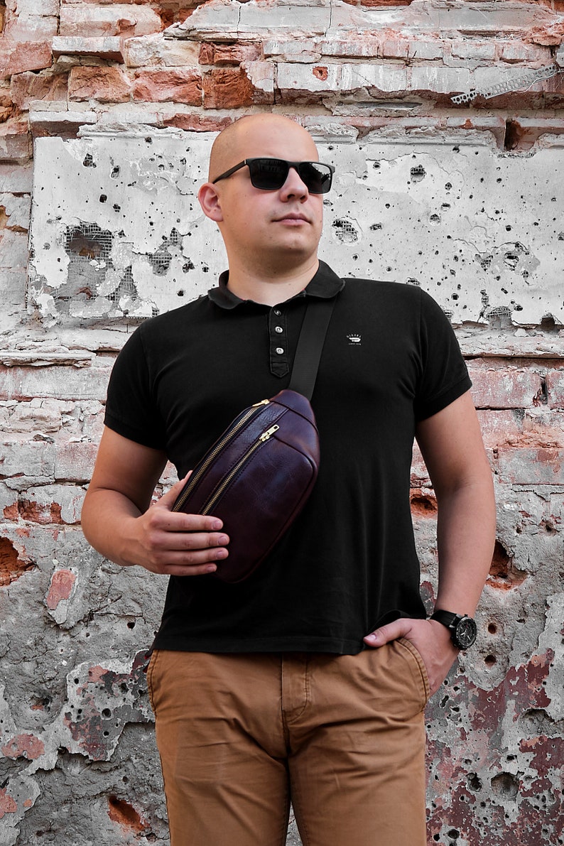 May include: A person wearing a black polo shirt with a brown leather fanny pack worn across the chest. The person is wearing brown trousers and sunglasses.