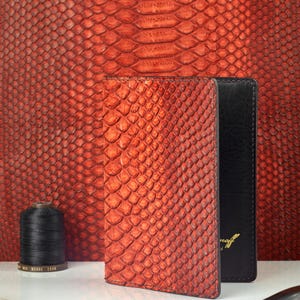 May include: A vertical wallet featuring a vibrant orange and red snakeskin-textured exterior with a black interior. The wallet is positioned upright, displaying its scale pattern and dark stitching, alongside a spool of black thread and metal tools.