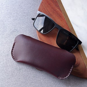 May include: A burgundy leather case for sunglasses. The case is closed and lying on a gray textured surface. A pair of black sunglasses is lying on a wooden surface in the background.