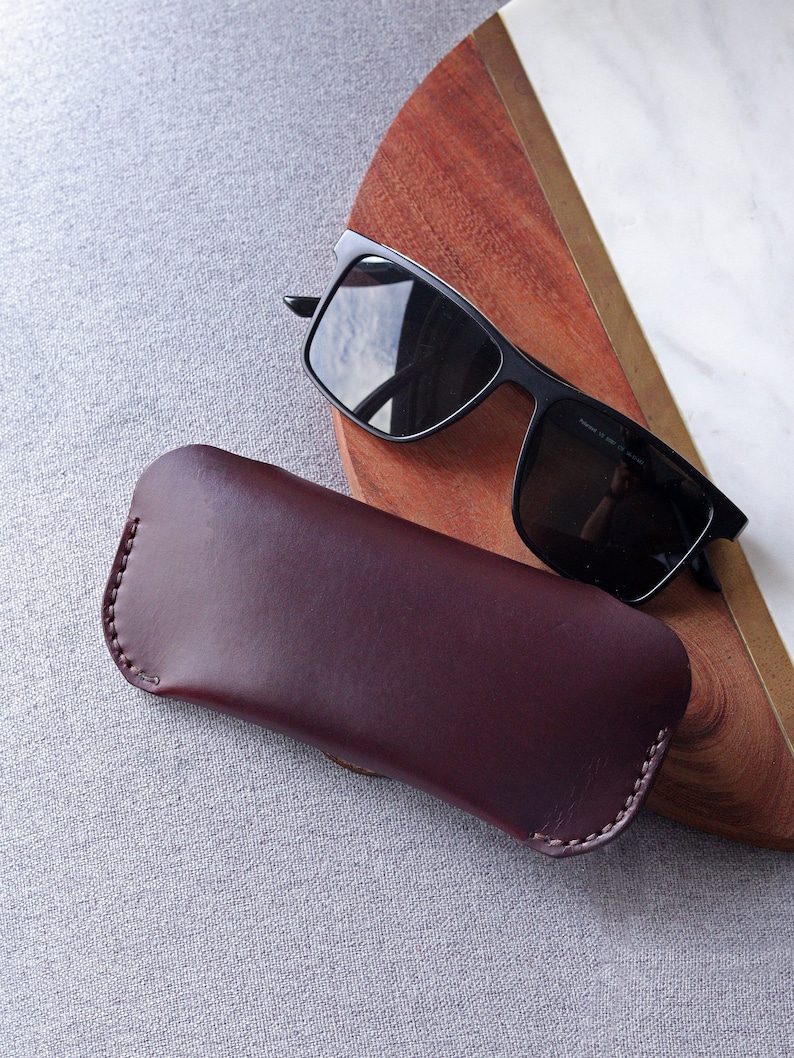 May include: A burgundy leather case for sunglasses. The case is closed and lying on a gray textured surface. A pair of black sunglasses is lying on a wooden surface in the background.