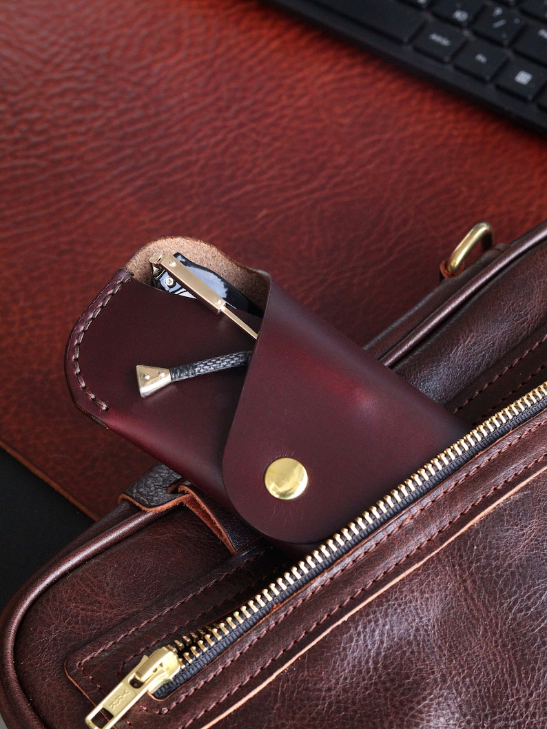 May include: A brown leather bag with a zipper closure. A burgundy leather glasses case with a gold button is partially inserted into the bag.