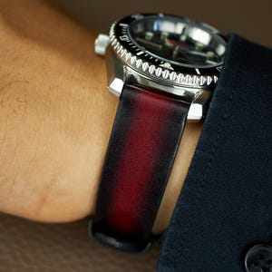 Puede incluir: Un reloj de pulsera negro y plateado con una correa de cuero roja. El reloj tiene una esfera negra con marcas blancas y un bisel giratorio.