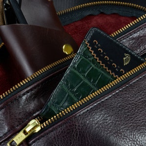 May include: A close-up of a brown leather bag with a zipper. The zipper is open and a green crocodile-embossed leather wallet is partially visible inside. The wallet has a black leather flap with a gold logo.