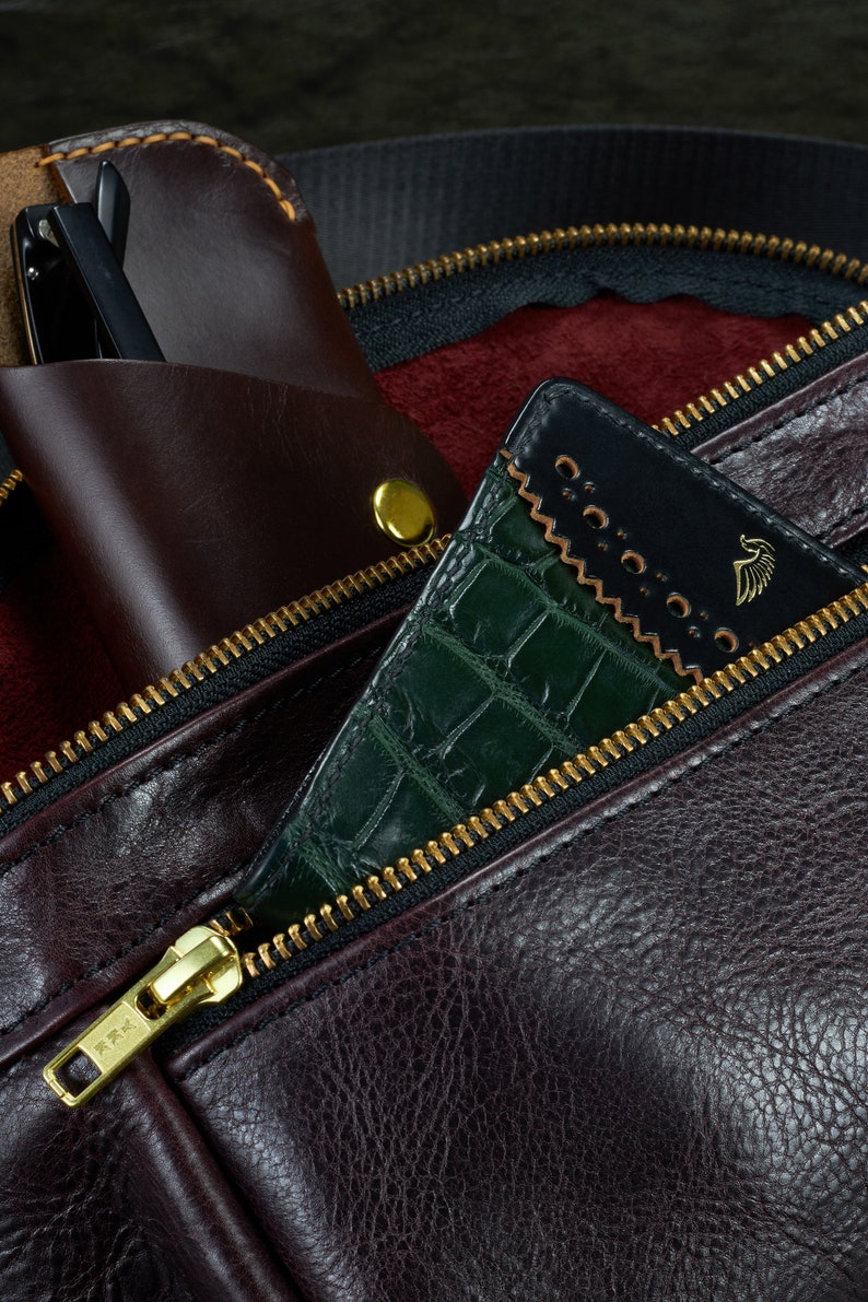 May include: A close-up of a brown leather bag with a zipper. The zipper is open and a green crocodile-embossed leather wallet is partially visible inside. The wallet has a black leather flap with a gold logo.