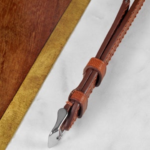 May include: A brown leather watch strap with a silver buckle. The strap has a stitched pattern and is shown on a white marble surface.