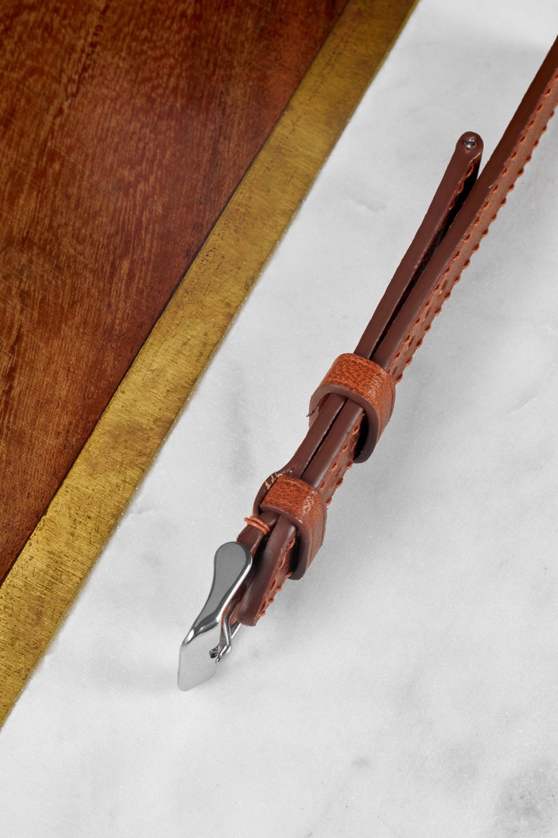 May include: A brown leather watch strap with a silver buckle. The strap has a stitched pattern and is shown on a white marble surface.