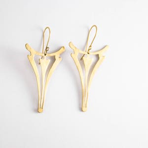 May include: A pair of gold-colored earrings with a unique, stylized design. Each earring features a central arrow-like shape with elongated, curved lines extending downwards. The earrings have a hook closure and are displayed against a white background.