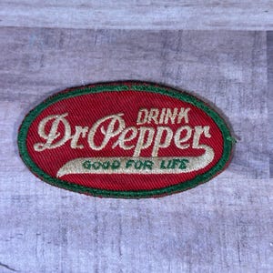 May include: An oval-shaped Dr. Pepper patch with a red background and a green border. The words "Dr. Pepper" are in a cursive font, with "DRINK" above and "GOOD FOR LIFE" below in a serif font.