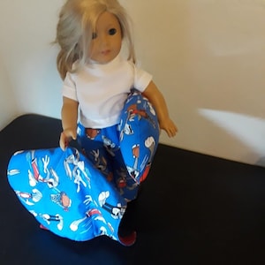 May include: A doll wearing a white shirt and blue pants with a cartoon character print. The doll is sitting on a black surface.