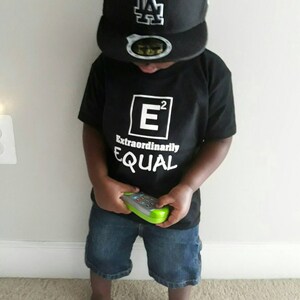 Può includere: Un bambino piccolo che indossa una maglietta nera con il testo "E² Extraordinarily EQUAL" e un berretto da baseball nero con un logo LA bianco. Il bambino tiene un giocattolo verde.
