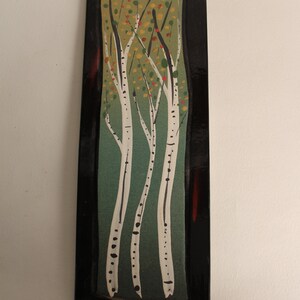 May include: A rectangular ceramic wall art piece with a black frame and a green background. The art features a stylized depiction of three birch trees with white bark and black markings. The trees are surrounded by small green and red leaves.