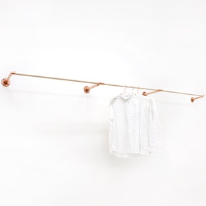 Könnte beinhalten: Ein Kleiderständer aus Kupferrohr mit zwei weißen Hemden, die daran hängen. Der Ständer ist an einer weißen Wand befestigt.