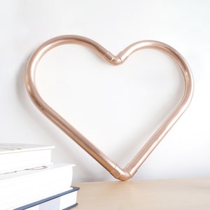May include: A rose gold heart-shaped metal wall decor with a smooth finish. The heart is hanging on a white wall and there is a vase of pink flowers on a wooden table below it.