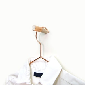 Puede incluir: Una camisa blanca con cuello colgada de una percha de oro rosa con un pomo de madera. La percha está fijada a una pared blanca.