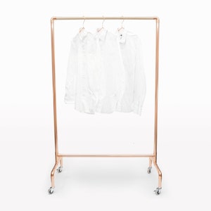 Pode incluir: Um cabideiro cor de cobre com três camisas brancas penduradas. O cabideiro tem uma estrutura retangular e assenta em quatro rodas. As camisas são brancas e limpas.