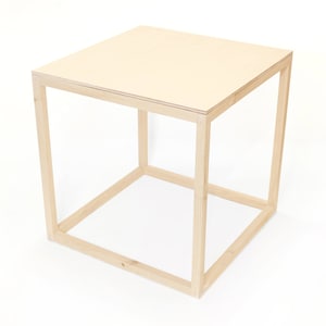 May include: A simple, light wood end table with a square top and a frame made of four pieces of wood. The table is made of natural wood and has a light, natural finish.
