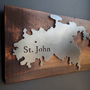 May include: A metal cutout of the island of St. John, mounted on a dark wood plaque. The island is outlined in silver metal and the name "St. John" is engraved in the metal.