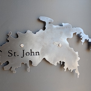 May include: A metal cutout of the island of St. John, with the name "St. John" inscribed on the island. The cutout is mounted on a gray wall.
