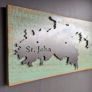 May include: A metal cutout of the island of St. John, mounted on a rustic wooden plaque with a distressed green paint finish. The island's name is inscribed on the metal cutout.