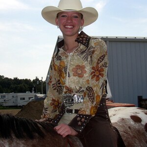 May include: A woman wearing a white cowboy hat with a brown and gold floral print shirt and brown pants riding a brown and white horse.