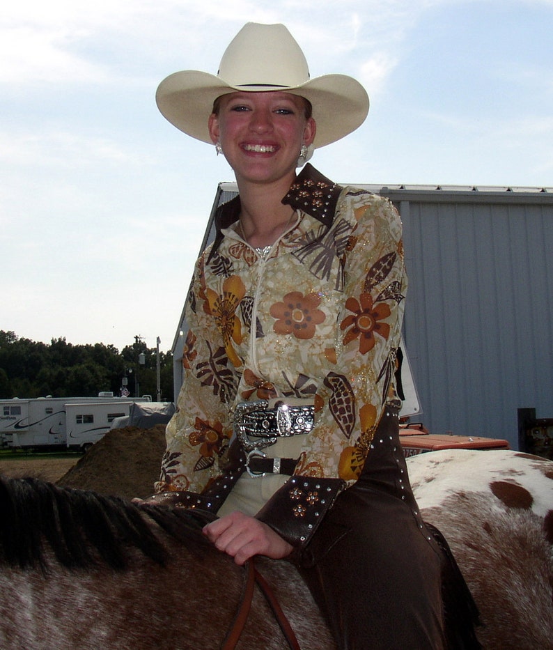 May include: A woman wearing a white cowboy hat with a brown and gold floral print shirt and brown pants riding a brown and white horse.