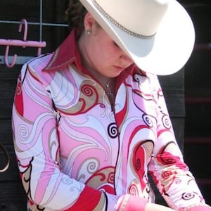May include: A woman wearing a pink, white, and brown patterned long-sleeved shirt with a white cowboy hat. She is wearing a black belt with rhinestones.