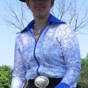 May include: A woman wearing a blue and white patterned shirt with a black cowboy hat and sunglasses. She is holding a brown leather saddle and has a silver belt buckle.
