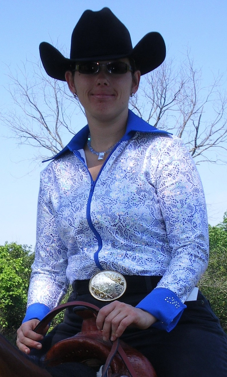 May include: A woman wearing a blue and white patterned shirt with a black cowboy hat and sunglasses. She is holding a brown leather saddle and has a silver belt buckle.