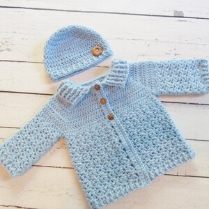 May include: A light blue crocheted baby cardigan with a matching hat. The cardigan has a collar and buttons down the front. The hat has a wooden button on top.