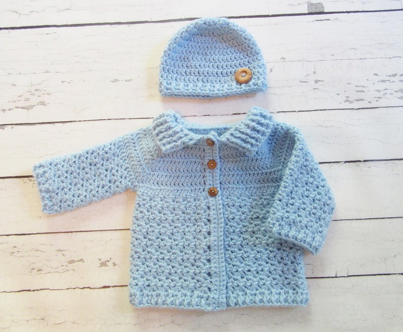 May include: A light blue crocheted baby cardigan with a matching hat. The cardigan has a button closure and a collar. The hat has a wooden button.