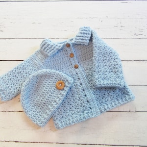 May include: A light blue crocheted baby cardigan with a matching hat. The cardigan has a collar and buttons down the front. The hat has a wooden button on top.