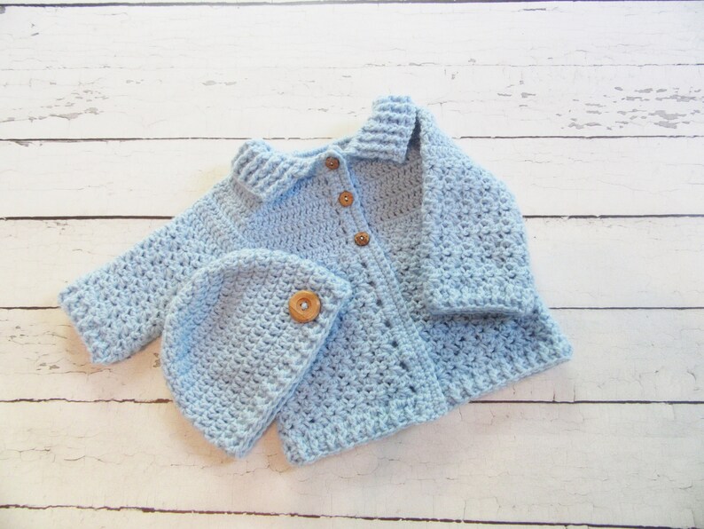 May include: A light blue crocheted baby cardigan with a matching hat. The cardigan has a collar and buttons down the front. The hat has a wooden button on top.