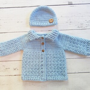 May include: A light blue crocheted baby cardigan with a matching hat. The cardigan has a collar and buttons down the front. The hat has a button on top.