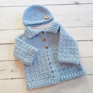 May include: A light blue crocheted baby cardigan with a matching hat. The cardigan has a collar and button closure. The hat has a wooden button.