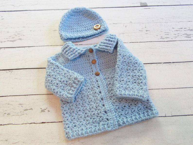 May include: A light blue crocheted baby cardigan with a matching hat. The cardigan has a collar and button closure. The hat has a wooden button.