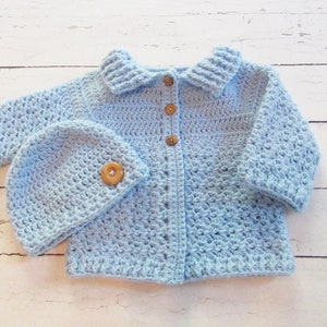May include: A light blue crocheted baby cardigan with a matching hat. The cardigan has a collar and buttons down the front. The hat has a simple design.