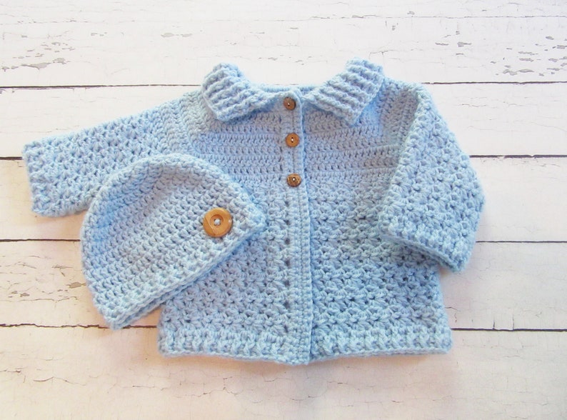 May include: A light blue crocheted baby cardigan with a matching hat. The cardigan has a collar and buttons down the front. The hat has a simple design.