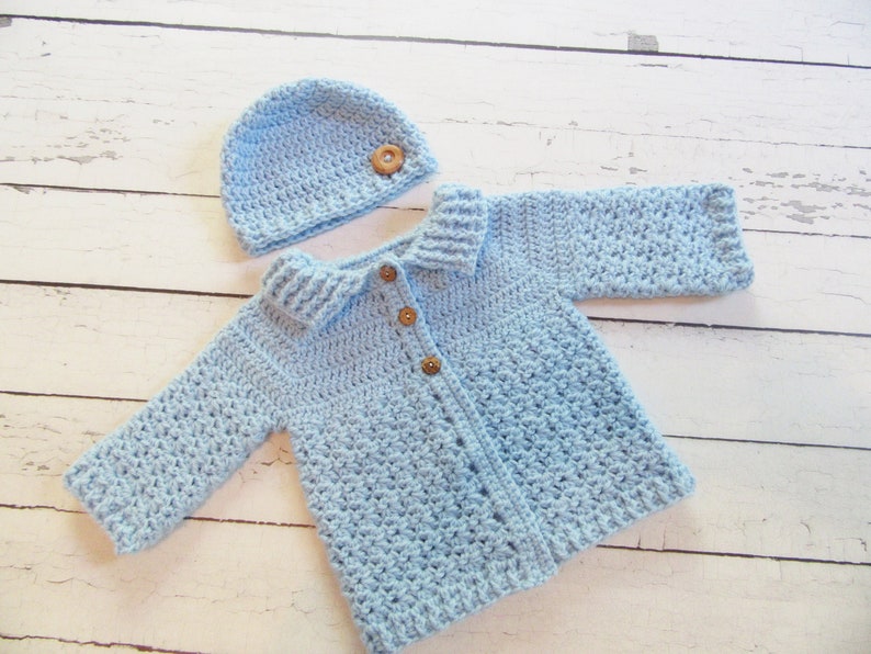 May include: A light blue crocheted baby cardigan with a matching hat. The cardigan has a collar and buttons down the front. The hat has a wooden button on top.