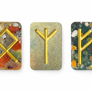 Peut inclure: Trois pierres de runes rectangulaires avec des symboles dorés. Les pierres ont un aspect moucheté avec des couleurs rouge, vert et or. Les runes sont de couleur or.