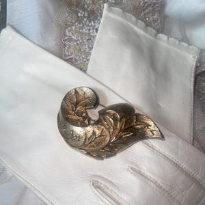 May include: A vintage gold-toned metal brooch with a leaf design, resting on a white leather glove. The brooch features intricate detailing and a classic aesthetic. The background includes white fabric and lace.